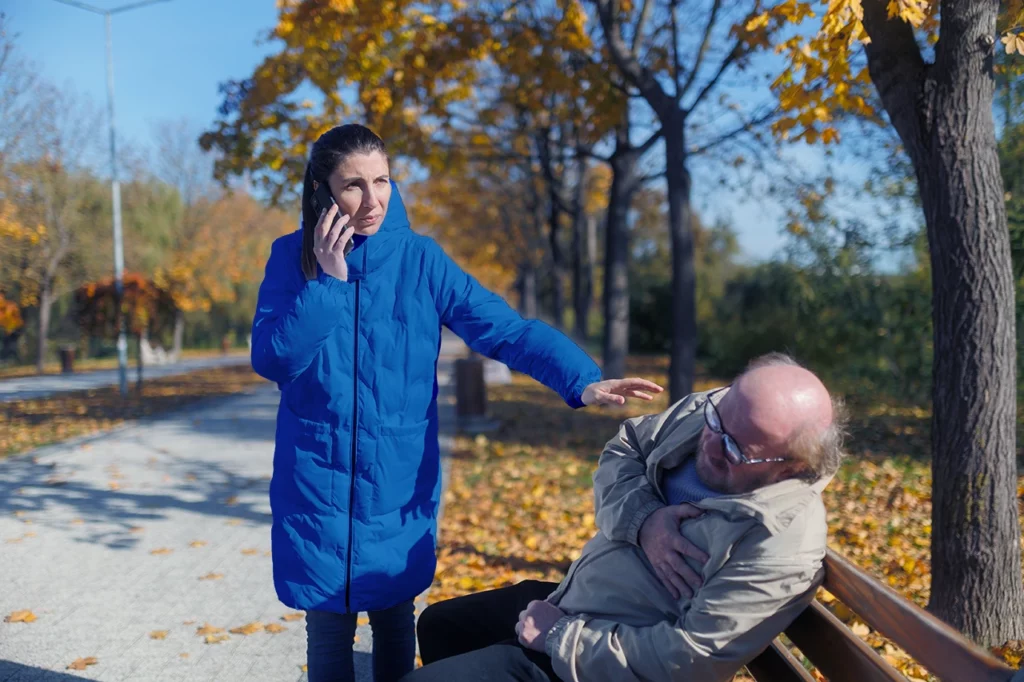 Mujer que sabe qué hacer en caso de infarto, llama por teléfono para pedir asistencia, mientras un hombre se infarta en un parque.