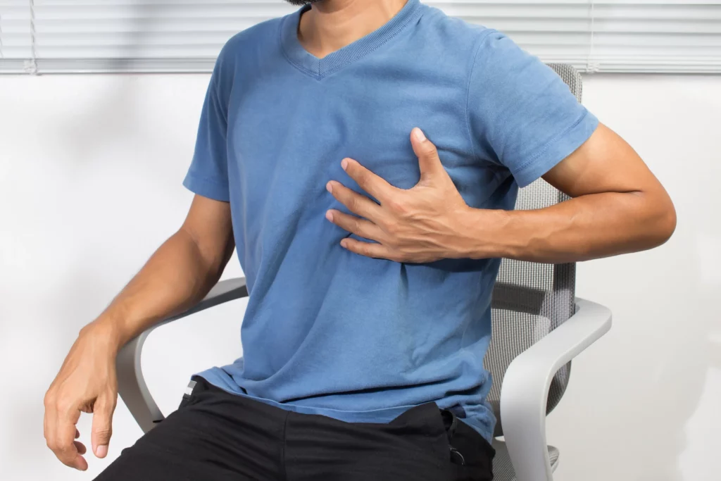 Hombre con angina de pecho y síntomas como dolor en el pecho, se encuentra sentado en una silla mientras espera atención médica.
