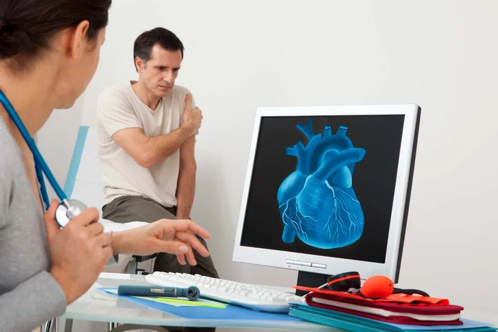 Hombre agarrándose el brazo debido a una condición qué es isquemia, mientras una doctora revisa una imagen del corazón.
