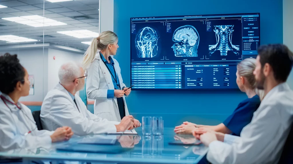 Médicos, del Tumor Board, en una sala de reuniones revisando los datos de un paciente en un monitor con radiografías de cabeza y cuello.
